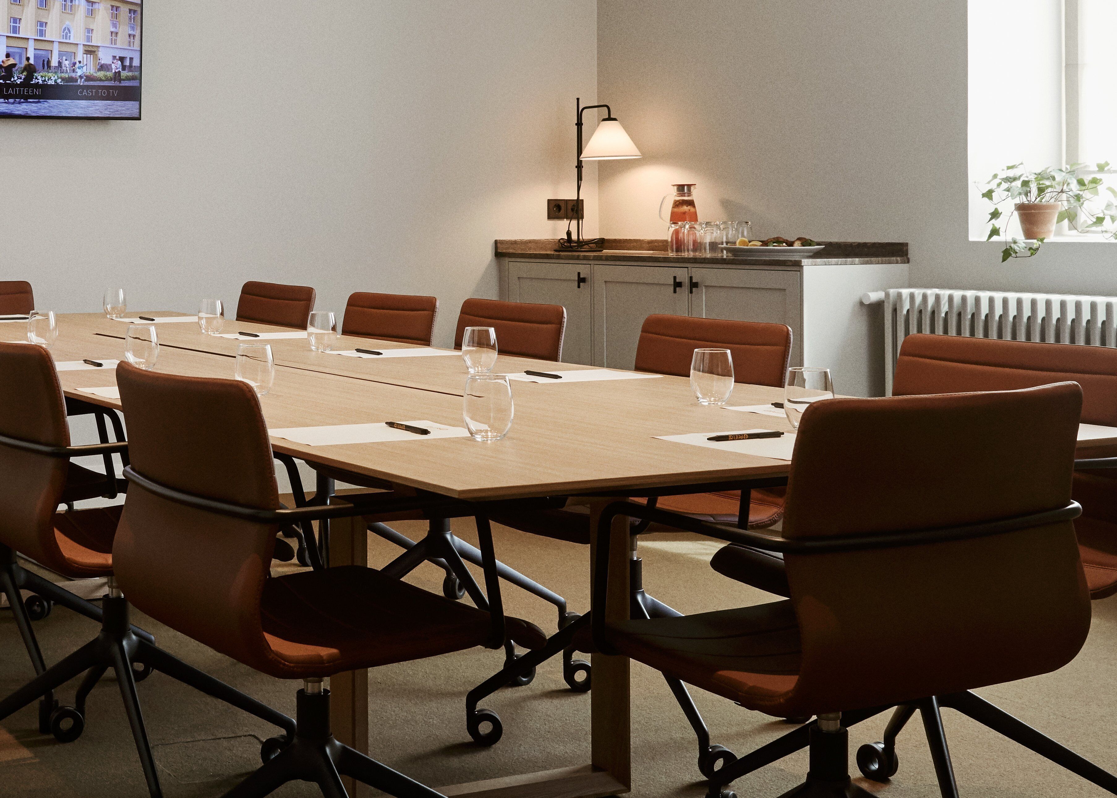 Een vergaderruimte met een lange tafel, bruine stoelen, een lamp en een radiator.