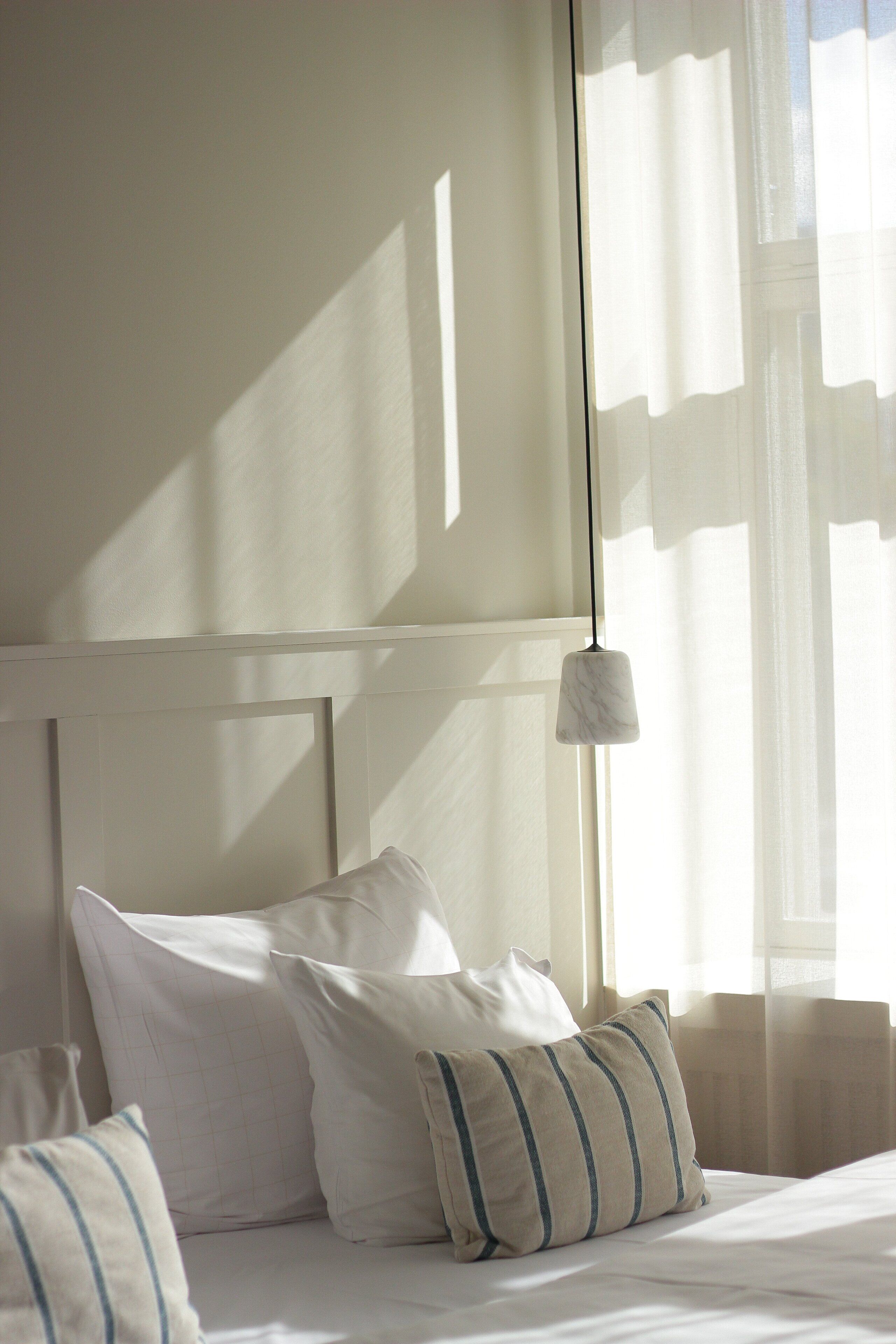 A neatly made bed with white bedding and striped pillows, a white headboard, and a hanging light fixture.