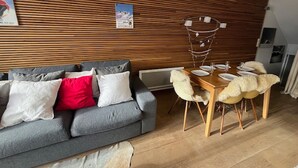 A modern living room with a grey sofa, wooden panel wall, and a dining area with a wooden table and chairs.