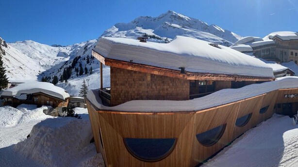 A wooden building with a sloped roof, surrounded by snow-covered mountains.