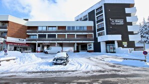 A multi-story building with a 'Sherpa' supermarket on the ground floor, a 'Maison du Tourisme' sign, and a snow-covered street.