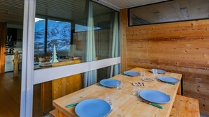 Un espace repas moderne avec une table en bois dressée pour quatre personnes, une cuisine visible à travers une porte vitrée et une vue sur un paysage montagneux enneigé.