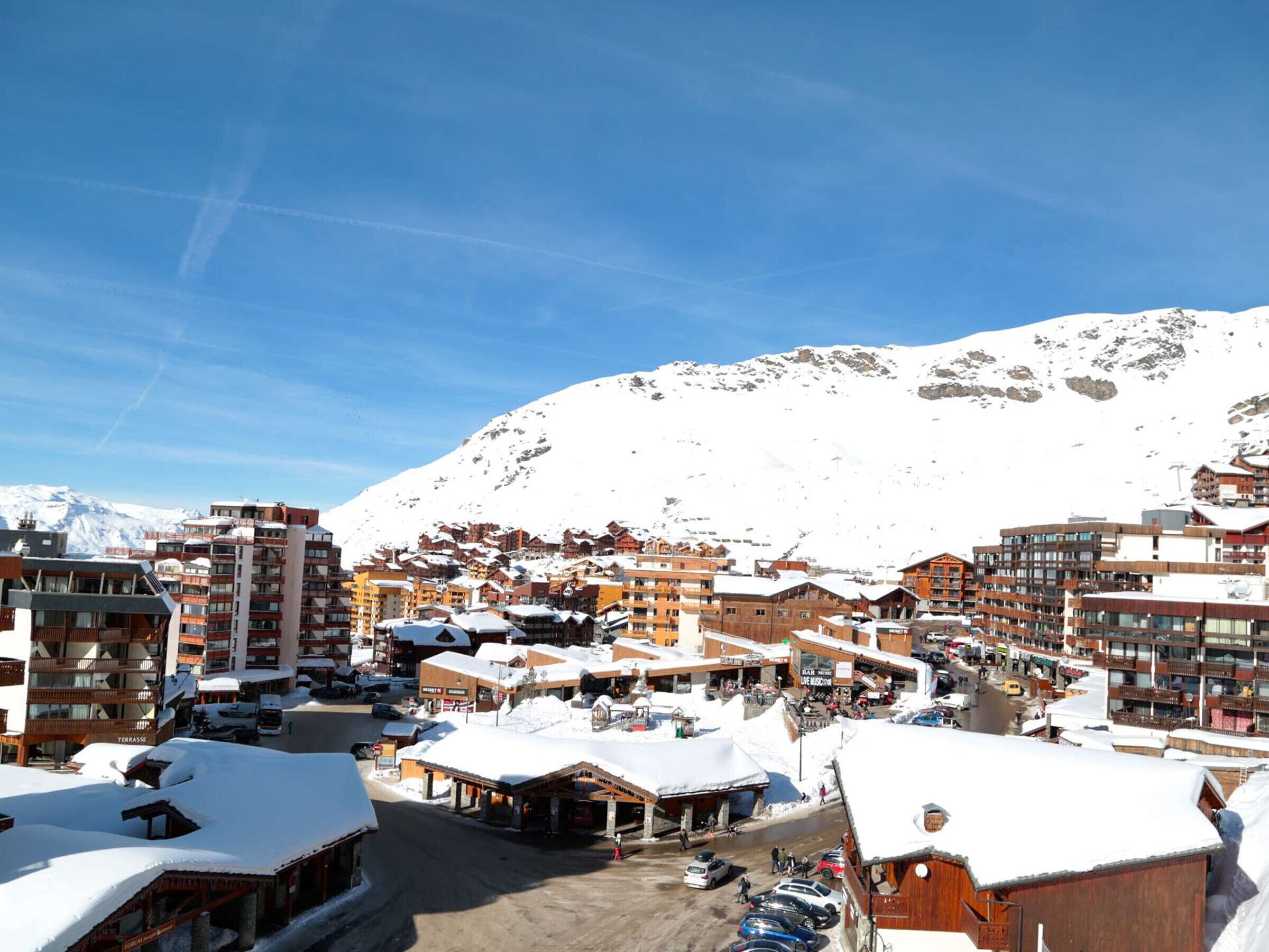 Un pueblo nevado en la montaña con varios edificios, incluyendo hoteles y tiendas, bajo un cielo azul despejado.