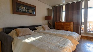 Une chambre avec deux lits, une commode en bois, un tableau et une fenêtre avec des rideaux.