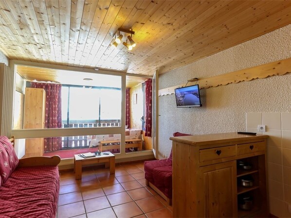 A cozy room with a wooden ceiling, a television mounted on the wall, a sofa, a small table, and a cabinet with shelves.