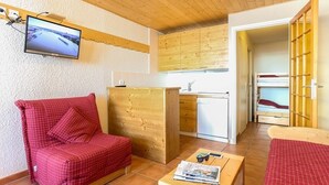 Ein kompakter Raum mit einem rot karierten Sofa, einem Holztisch mit Zeitschriften, einem an der Wand befestigten Fernseher, einer Küchenzeile und einem Etagenbett.