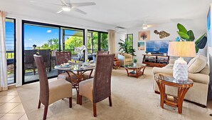 A spacious living room with a dining area, large windows, ceiling fans, and a television. - Tropical Escape with Endless Coastal Views (Kihei)