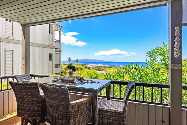 A balcony with wicker chairs and a glass table, overlooking a coastal landscape with greenery and a clear sky.