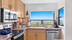 A kitchen with wooden cabinets, stainless steel appliances, a large window with a view, and a countertop with a kettle and coffee maker. - Tropical Escape with Endless Coastal Views (Kihei)