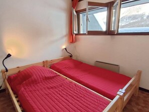 Um quarto pequeno com duas camas de madeira, cada uma com colchão vermelho. Há uma janela com cortinas, uma luminária fixada na parede e um radiador.
