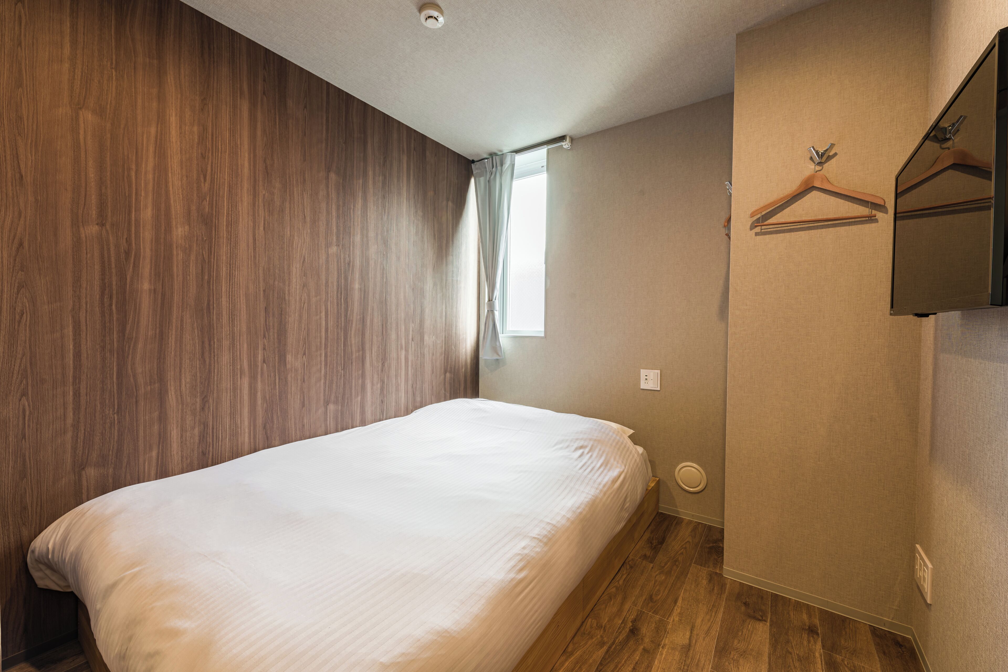 A bedroom with a wooden headboard, a single bed, a window with a curtain, and a wall-mounted mirror with a wooden hanger.