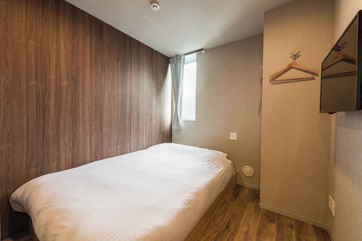 A bedroom with a wooden headboard, a single bed, a window with a curtain, and a wall-mounted mirror with a wooden hanger.