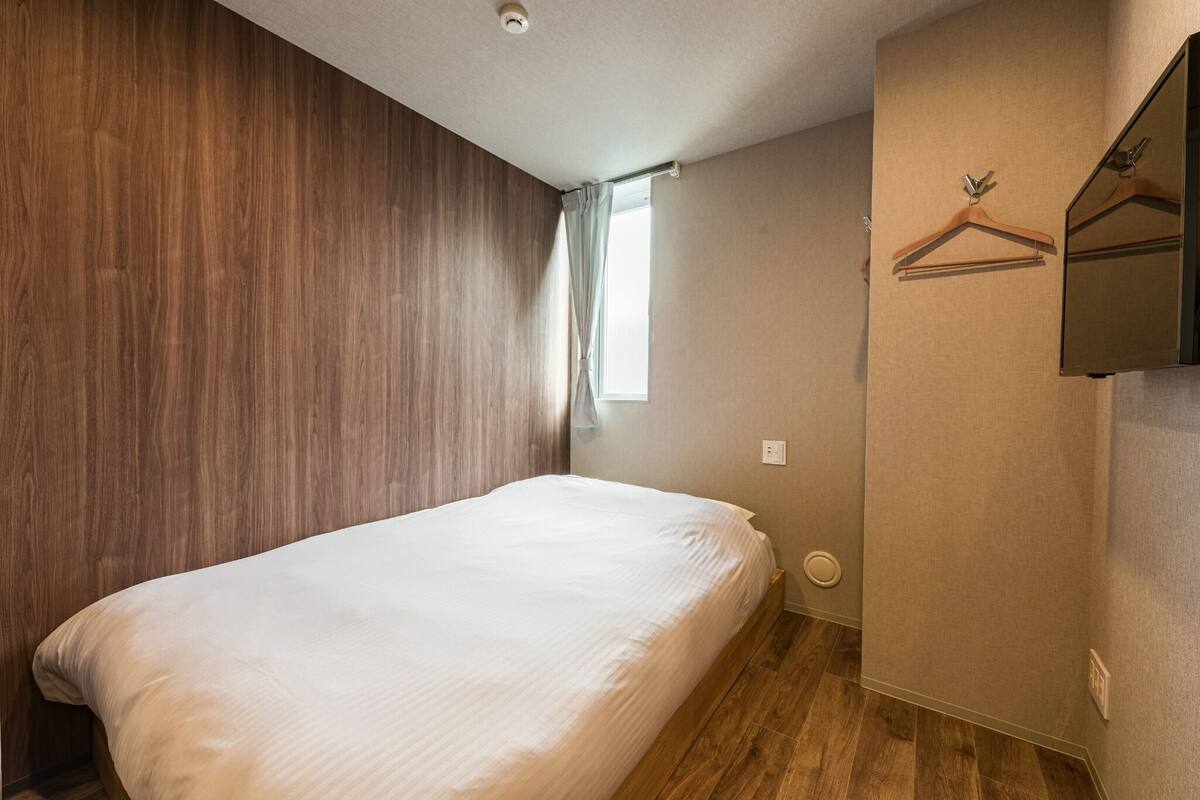 A bedroom with a wooden headboard, a single bed, a window with a curtain, a wall-mounted mirror, and a wooden floor.