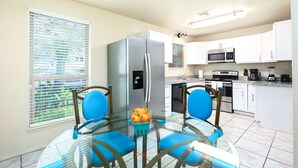 A modern kitchen with white cabinets, a stainless steel refrigerator, and a glass dining table with blue chairs.