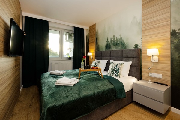 A cosy bedroom combining the naturalness of wood with a wallpaper depicting a misty forest. A flat-screen TV hangs on the wall and dark curtains help to darken the room.