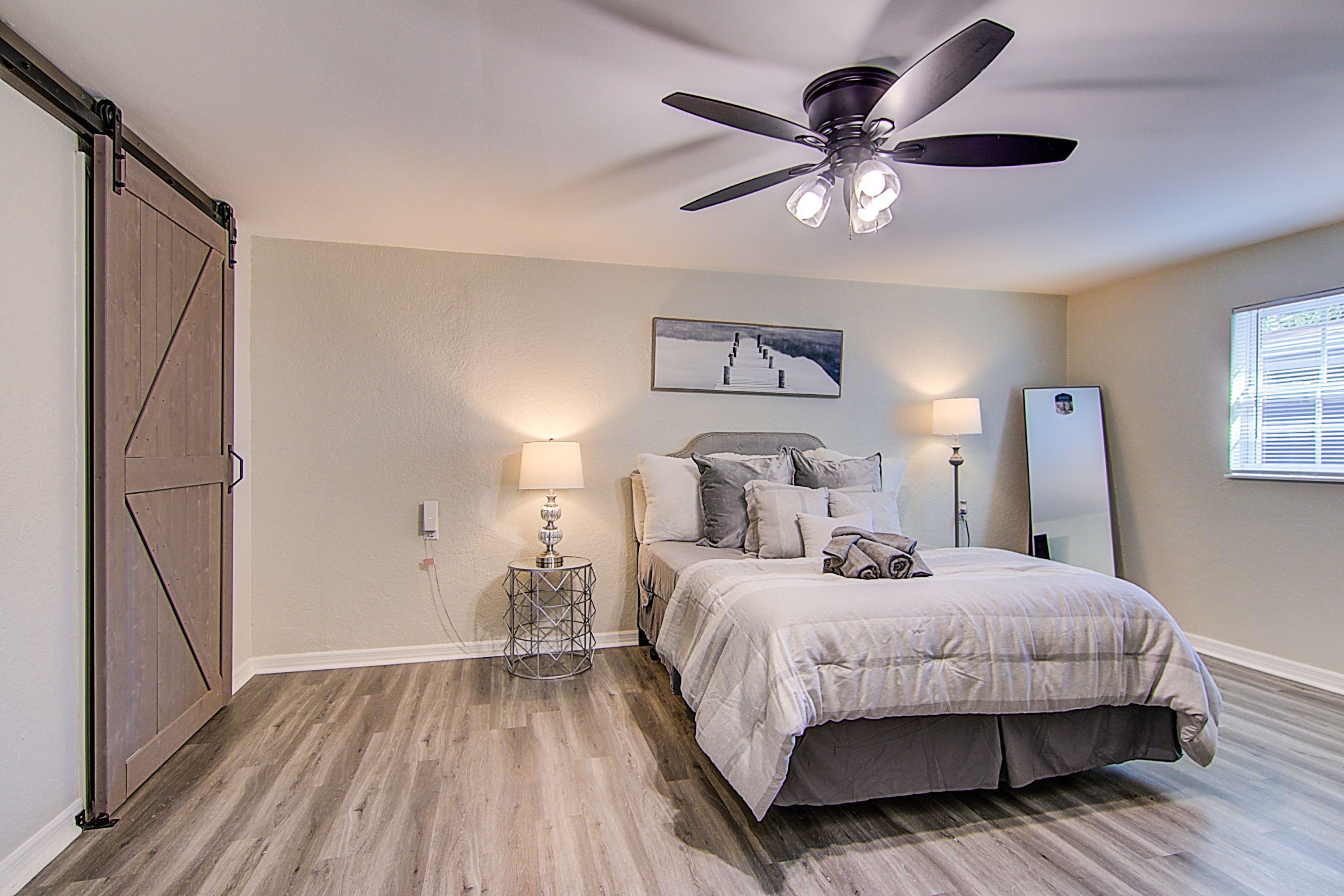 A bedroom with a bed, a ceiling fan, a nightstand, a lamp, a picture on the wall, and a window with blinds.