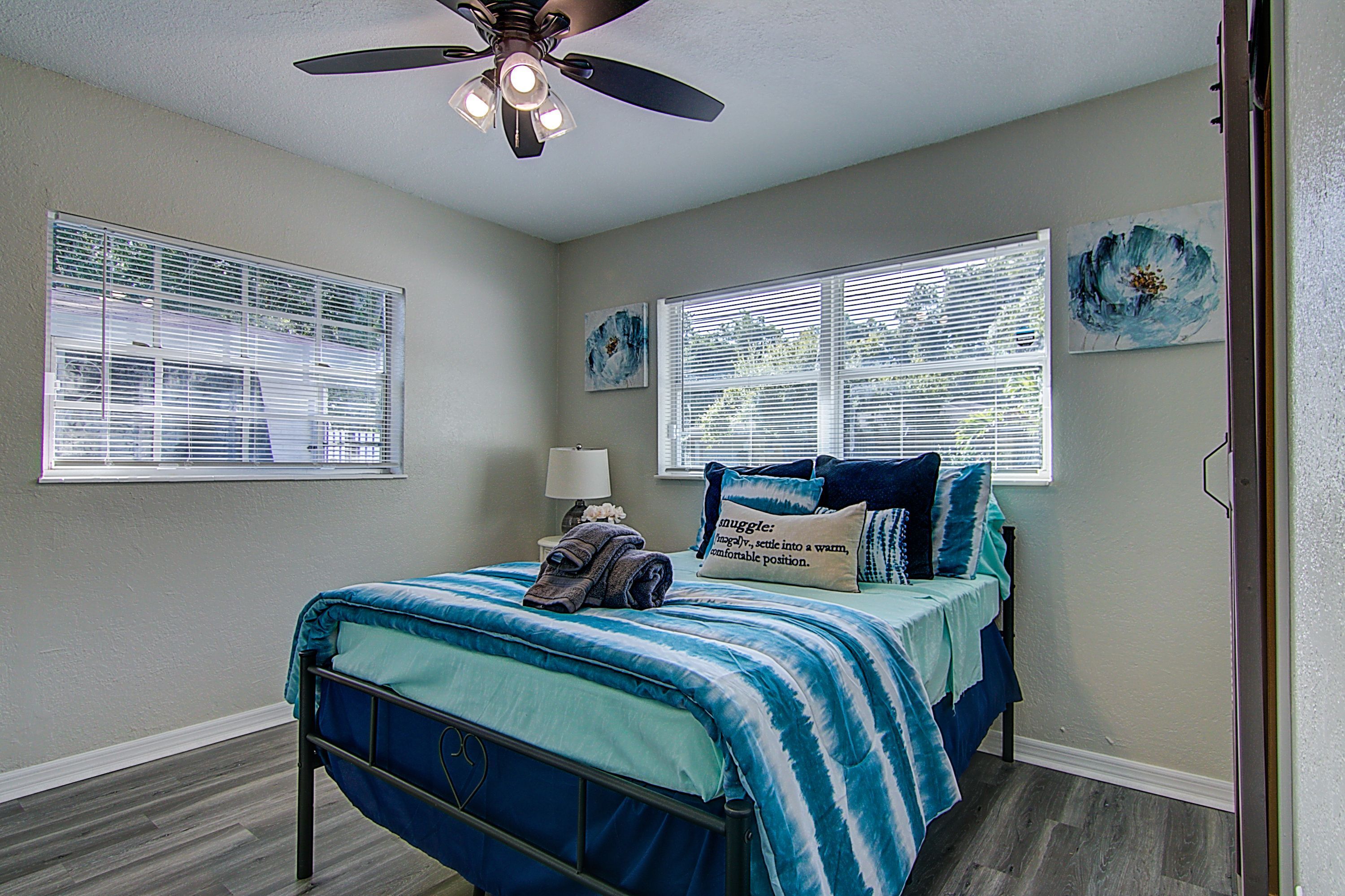 A bedroom with a bed, a ceiling fan, and two windows with blinds.