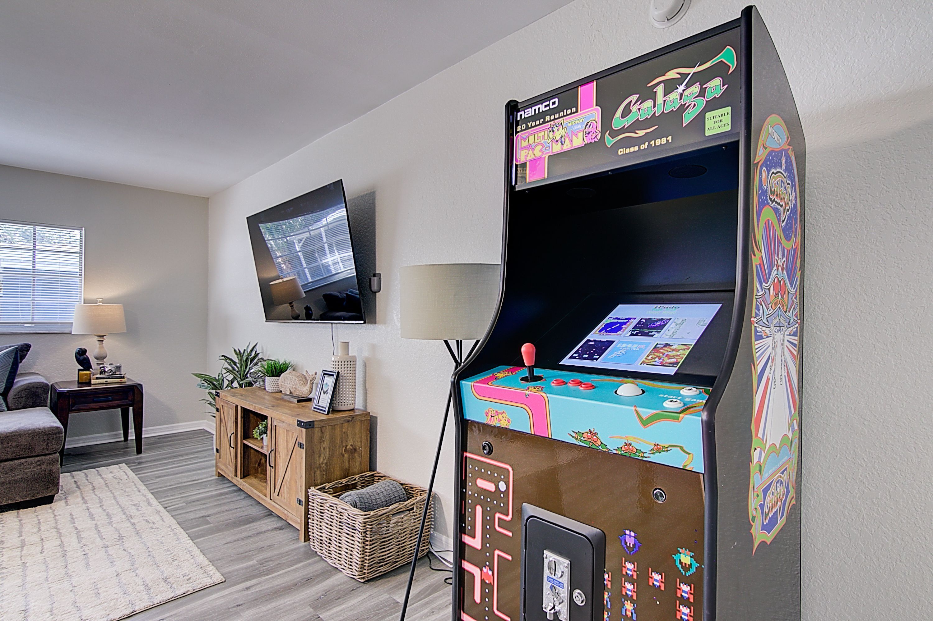 Una sala de estar con un televisor de pantalla plana grande montado en la pared, un mueble de madera, una cesta de mimbre y una máquina arcade colorida.