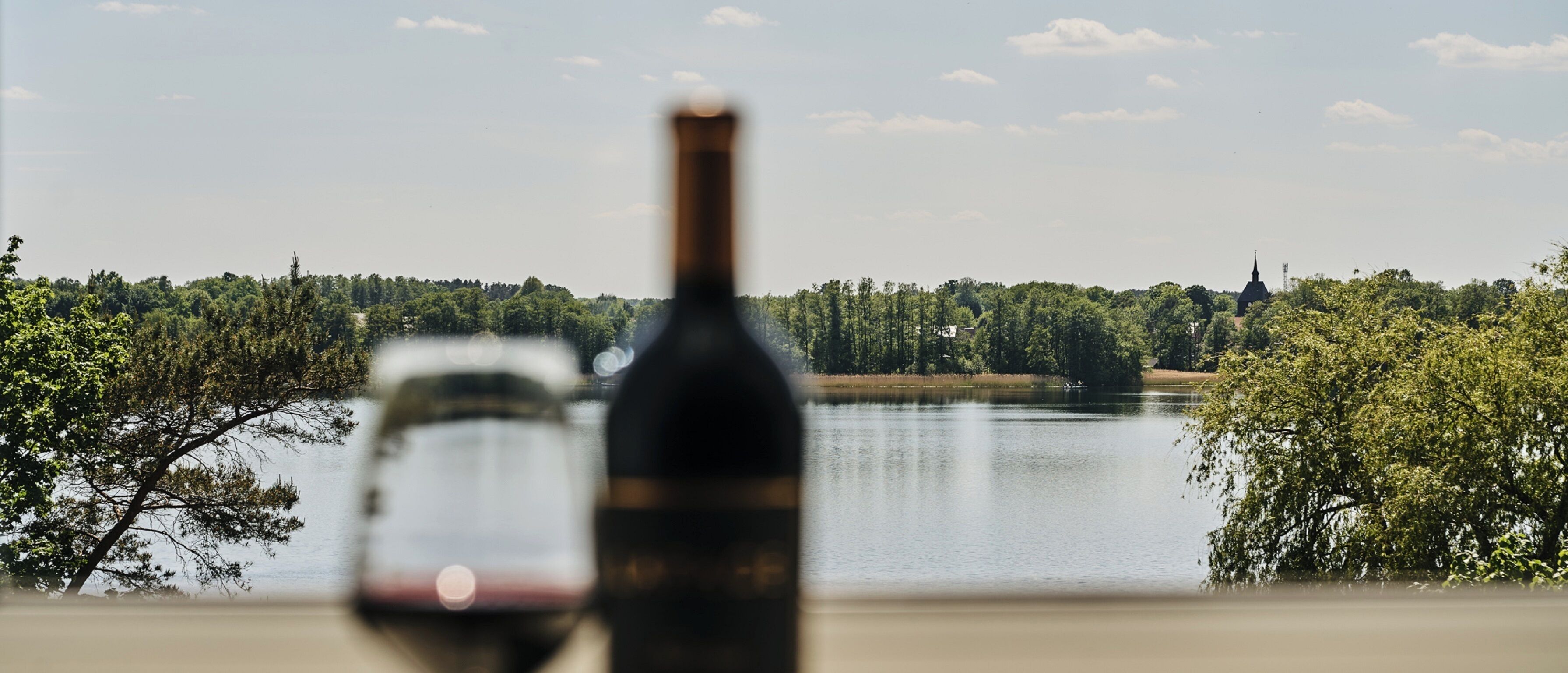 A bottle and glass of wine in the foreground with a lake and trees in the background.