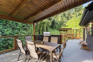 Une terrasse extérieure avec une table et des chaises en bois, un barbecue et une vue sur une zone boisée.