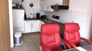 Une cuisine de taille réduite avec des meubles blancs, un four à micro-ondes, une machine à café et un évier. On y trouve deux chaises rouges et une poubelle.