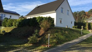 Une maison blanche avec un toit en pente, entourée de verdure et bordée d’un chemin pavé.