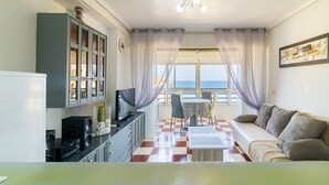 Un espace de vie compact comprenant un canapé, une table à manger et un coin cuisine. Une grande fenêtre offre une vue sur la mer.