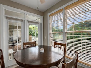 A dining area with a round table, four chairs, and a view through large windows with blinds.