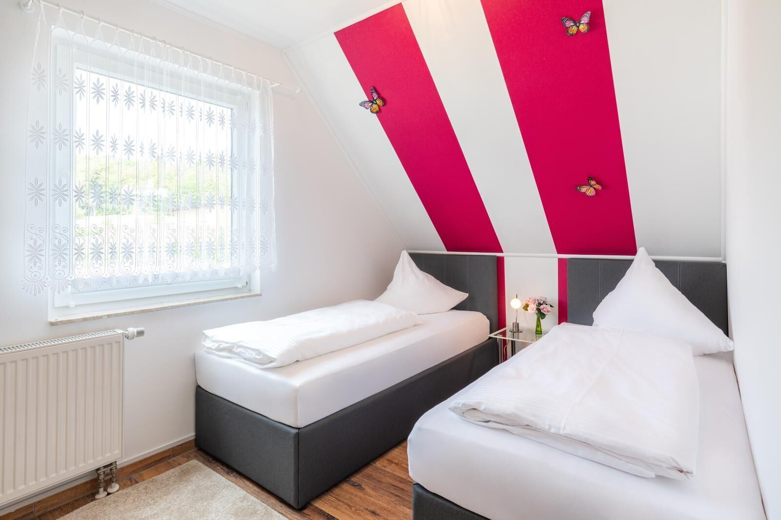 Une chambre avec deux lits, une fenêtre agrémentée de rideaux décoratifs, un radiateur et un miroir sur lequel est posé un vase de fleurs.