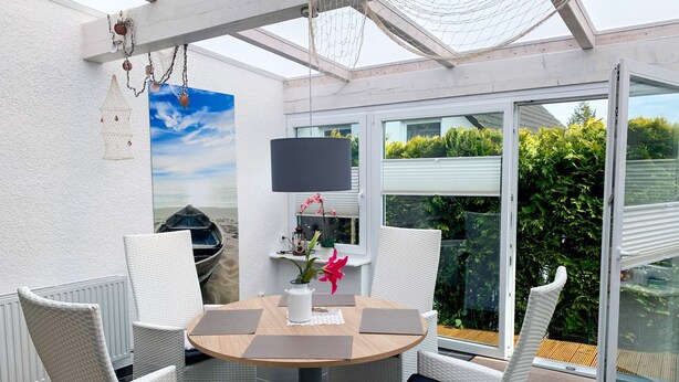 Eine sonnendurchflutete Terrasse mit einem runden Tisch, weißen Korbstühlen, einem Strandbild und Blick auf Grün hinter einer Glastür.