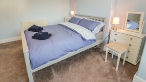 Una cama bien hecha con ropa doblada, una lámpara de noche, un pequeño banco de madera y un aparador con espejo.