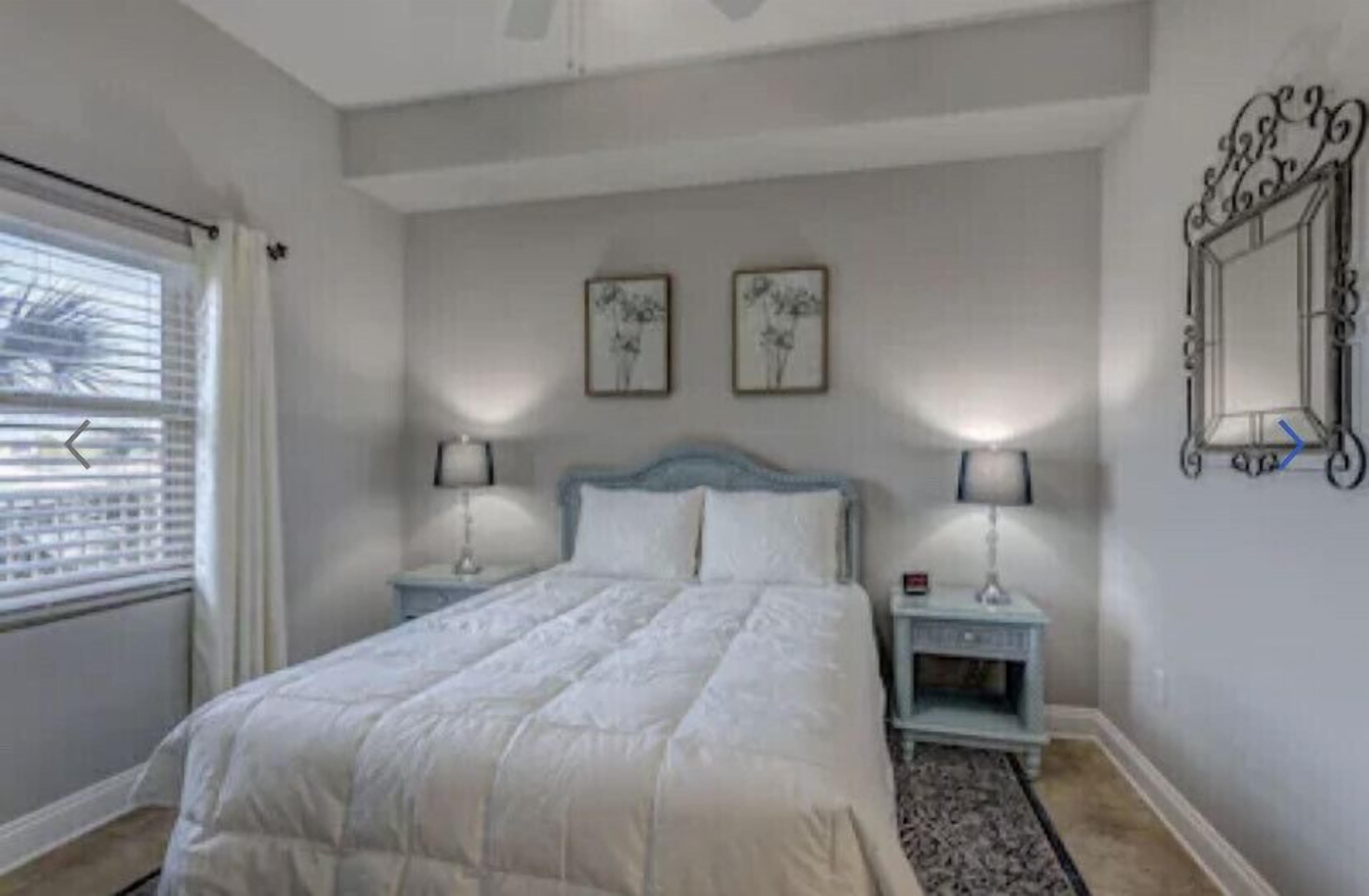 Un dormitorio con una cama, dos mesitas de noche, una lámpara de noche, una ventana con persianas y dos cuadros colgados en la pared.