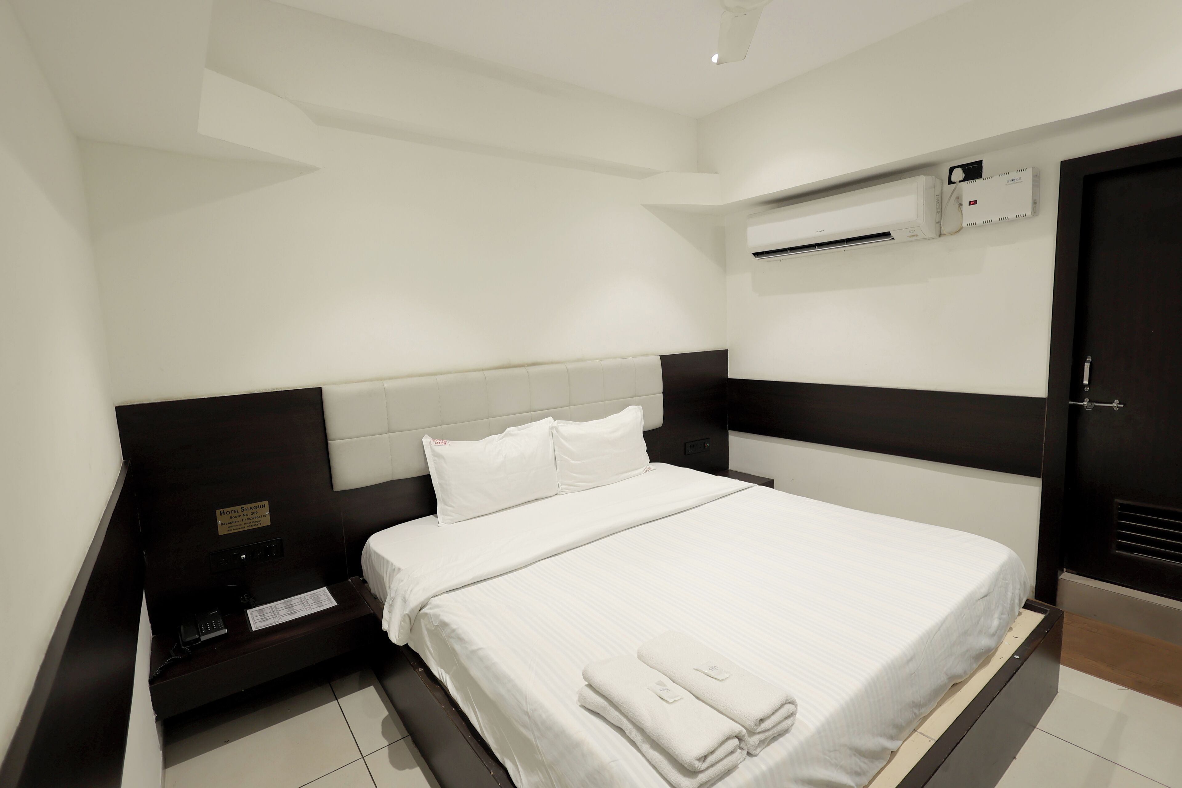 A hotel room with a large bed, a bedside table with a phone, a nightstand, and a wall-mounted air conditioning unit.