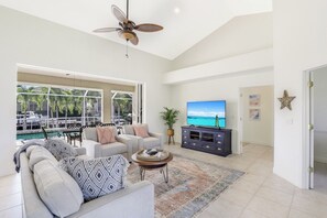 A spacious living room with a large TV, comfortable seating, a ceiling fan, and a view of the ocean through large windows.