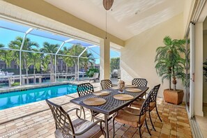 Uma área de refeições ao ar livre com vista para a piscina, incluindo uma mesa posta com jogos americanos e cadeiras, uma planta em vaso e uma churrasqueira.