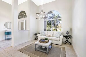 A bright living room with a white sofa, a coffee table, and a large window with a view of palm trees.