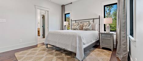 Un dormitorio con cama, mesita de noche, ventilador de techo, ventana con cortinas y puerta que da a otra habitación.