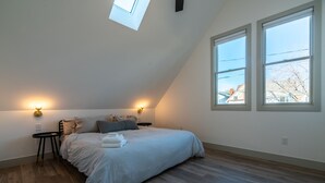 Ein Schlafzimmer mit schrägverdeckter Decke, einem Bett mit Kissen, zwei Nachttischlampen und einem Dachfenster.