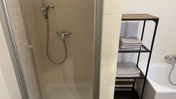 A bathroom with a glass-enclosed shower, a white bathtub, and a shelf with folded towels.