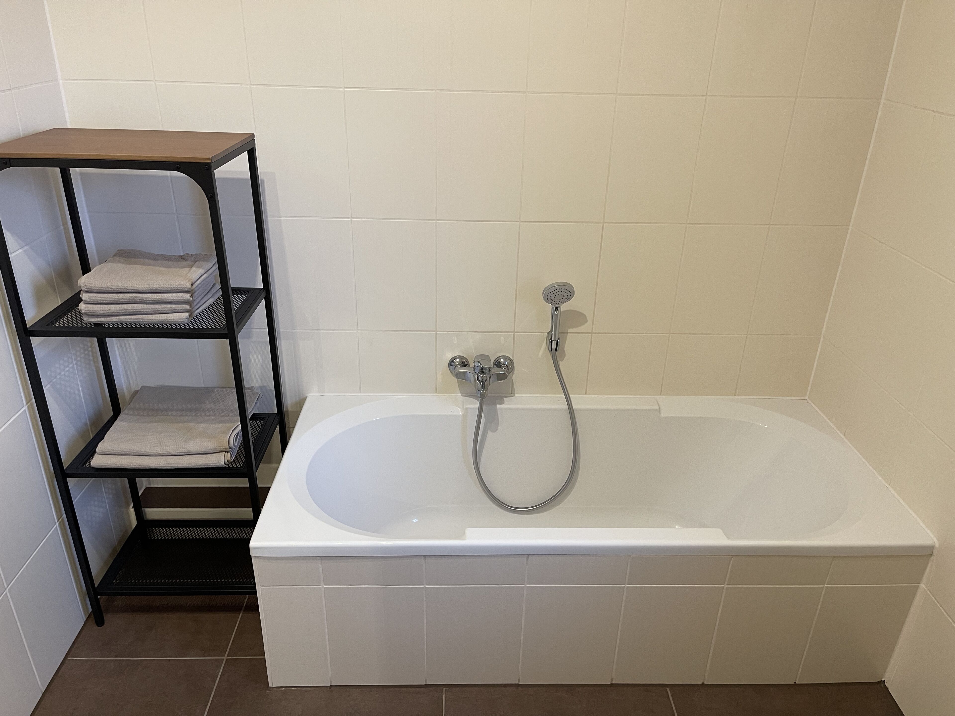 A bathroom with a white bathtub, a showerhead, and a black metal shelf with folded towels.