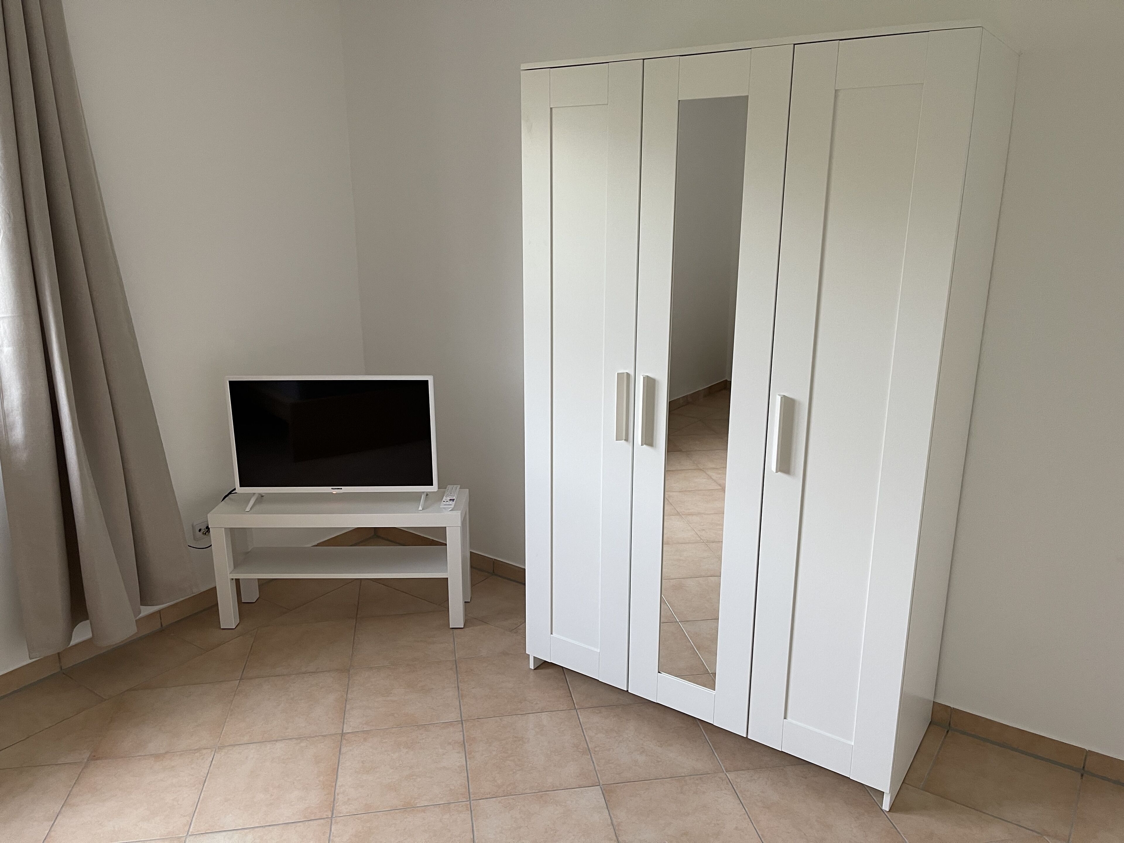 Una habitación con una televisión sobre una mesita, un armario con puertas espejadas y suelo de baldosas.