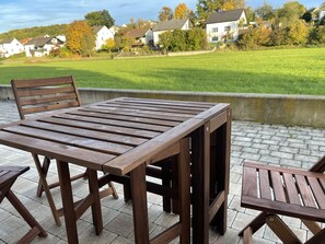 A wooden outdoor table and chairs on a paved patio with a view of houses and a green field in the background. - Apartment Mühlbach (Bad Abbach)