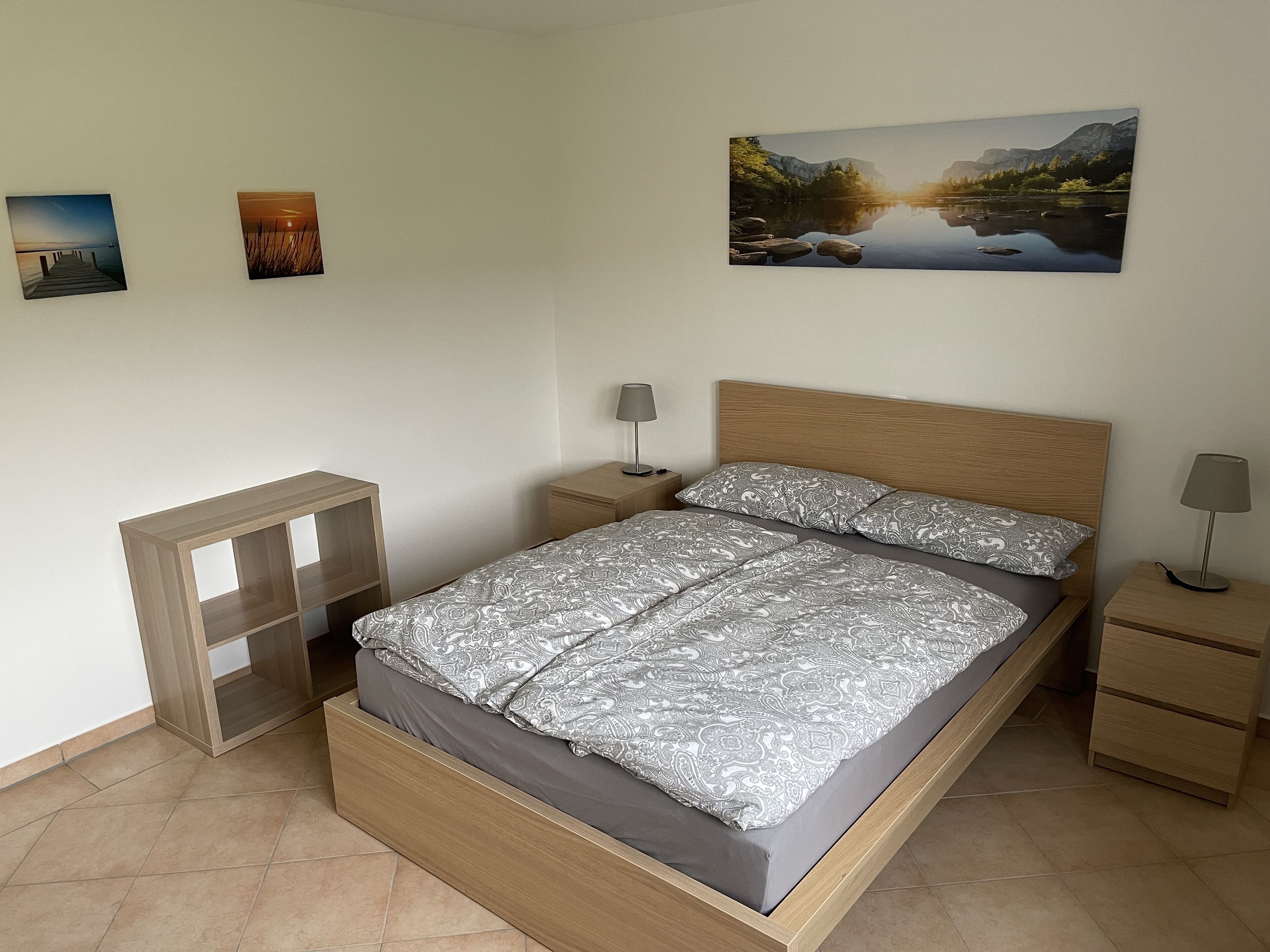 Un dormitorio con una cama de madera, una mesita de noche, un pequeño estante de madera y un cuadro de un paisaje en la pared.