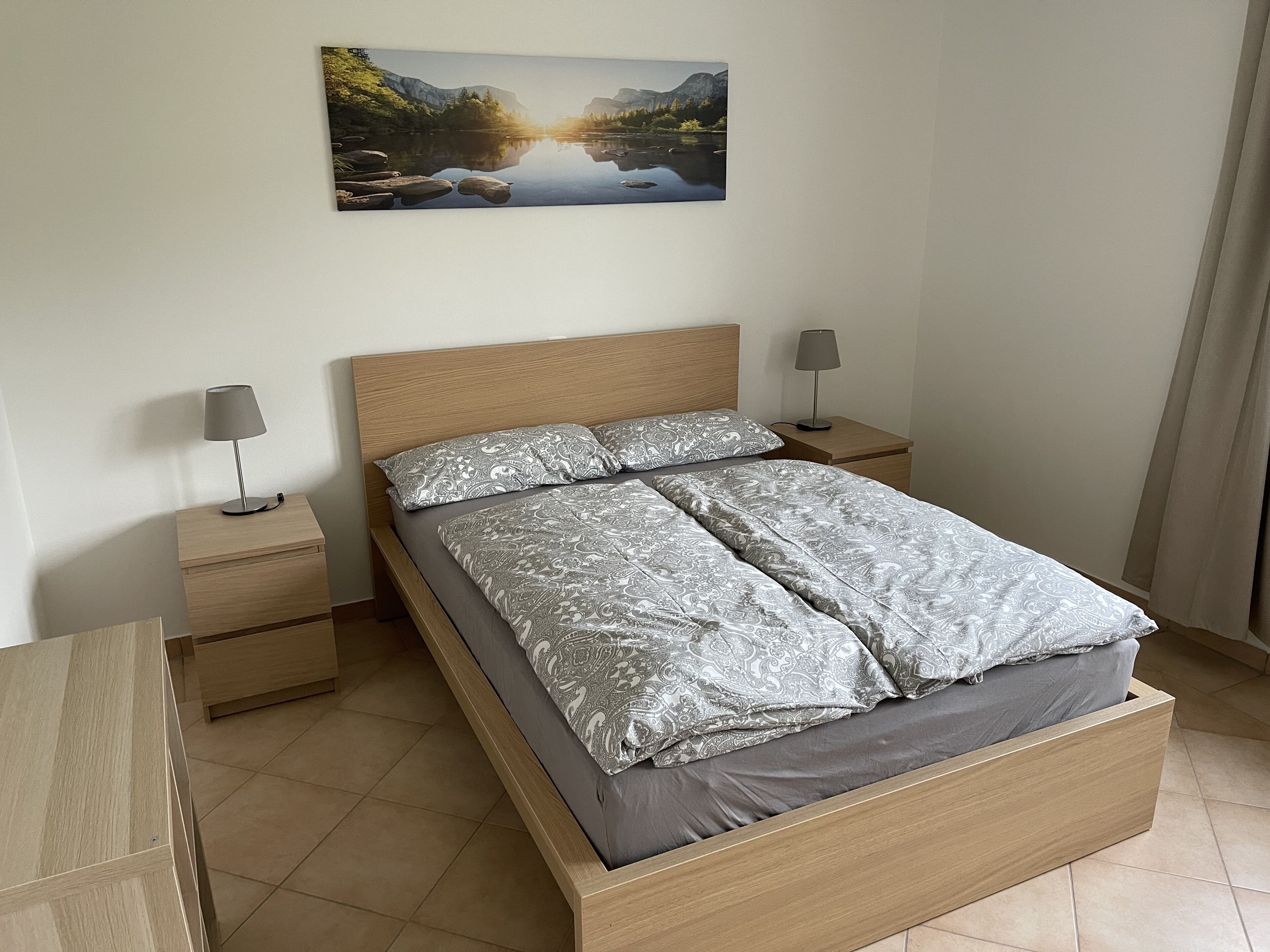Une chambre à coucher avec un lit en bois, deux tables de chevet, une lampe sur chacune, et un tableau représentant un paysage accroché au mur.