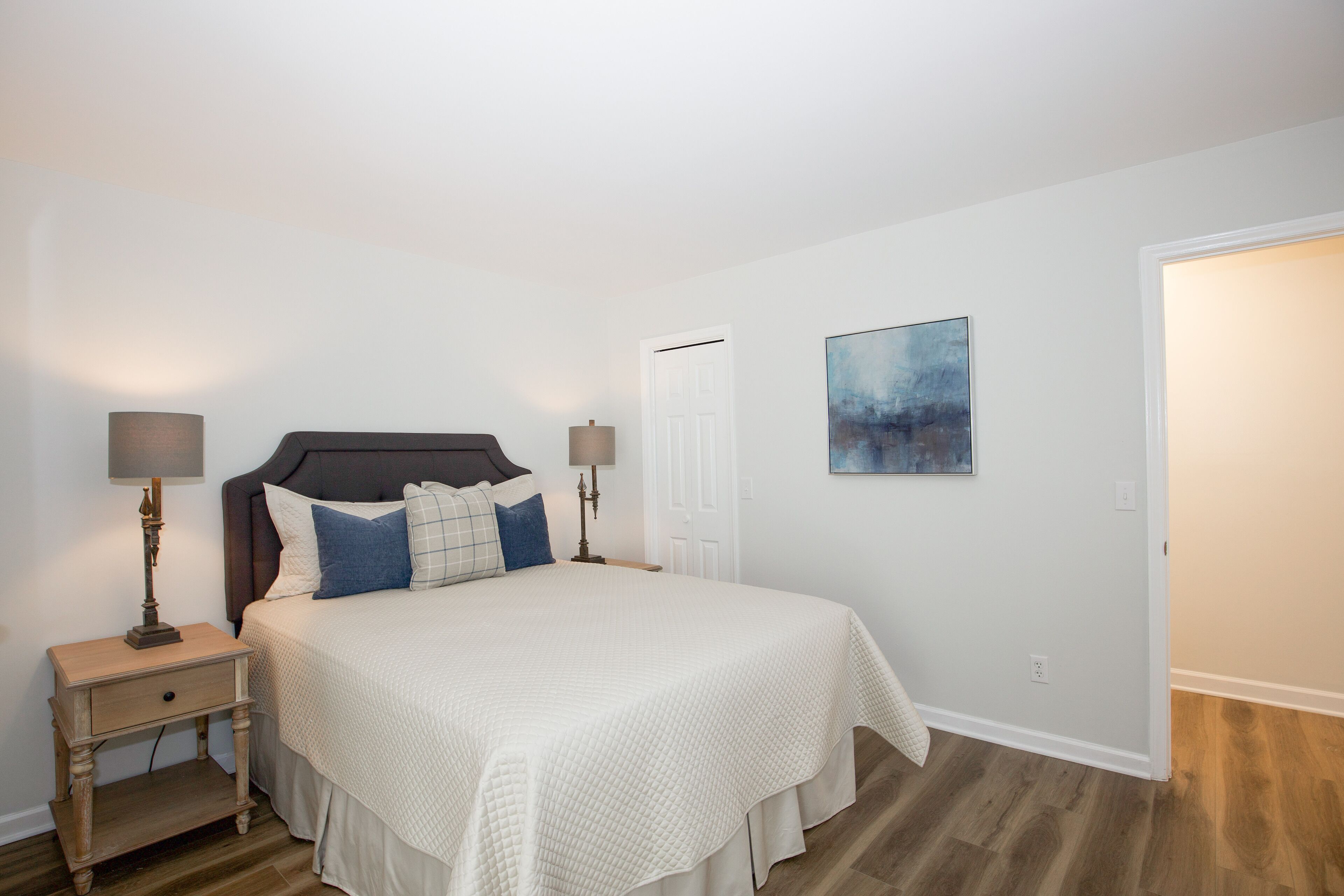 A bedroom with a large bed, two bedside tables with lamps, a door, and a painting on the wall.