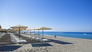 A beach with rows of white lounge chairs and beige umbrellas, a clear blue sky, and calm sea.