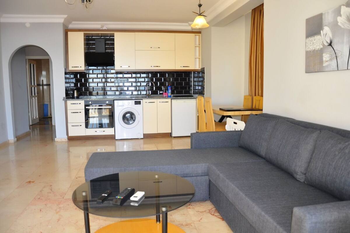 A modern apartment with a grey sofa, a glass coffee table, a washing machine, and a kitchen with black and white tiles.