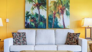Een lichtgrijze bank met gekleurde kussens, een glazen salontafel, en twee abstracte schilderijen van palmboompjes op gele muren.