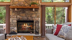 A stone fireplace with a fire, a wooden mantel, and a view of trees through large windows. - Pocono Pines Family Retreat: Game Room, Sunroom & Pets Welcome! (Pocono Pines)