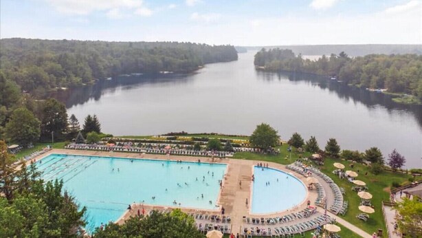 A large outdoor swimming pool surrounded by a deck with lounge chairs and umbrellas, set against a backdrop of a calm lake and forested hills. - Pocono Pines Family Retreat: Game Room, Sunroom & Pets Welcome! (Pocono Pines)
